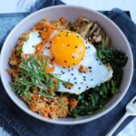 Overhead view of a bowl containing a fried egg topped with sesame seeds and drizzled with orange sauce, surrounded by quinoa, spinach, and mushrooms, on a metal tray with a fork and dark blue fabric.