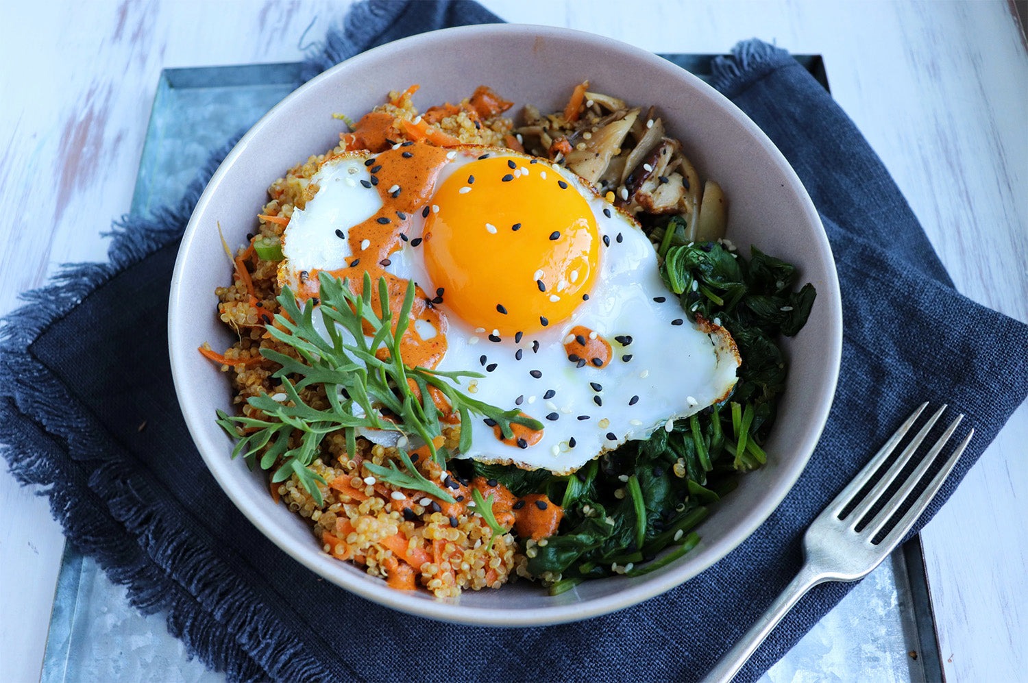 Overhead view of a bowl containing a fried egg topped with sesame seeds and drizzled with orange sauce, surrounded by quinoa, spinach, and mushrooms, on a metal tray with a fork and dark blue fabric.