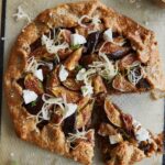 A rustic, partially sliced fig and goat cheese galette, with browned flaky crust, filled with caramelized onions and sliced fresh figs, garnished with dollops of white goat cheese, sauerkraut, fresh thyme, and large salt flakes on a parchment-lined baking sheet.