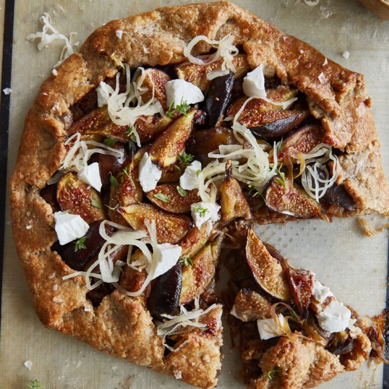 A rustic, partially sliced fig and goat cheese galette, with browned flaky crust, filled with caramelized onions and sliced fresh figs, garnished with dollops of white goat cheese, sauerkraut, fresh thyme, and large salt flakes on a parchment-lined baking sheet.