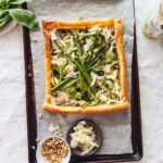 Overhead shot of a rustic tart on a baking sheet, featuring a golden-brown puff pastry crust filled with courgette ribbons, asparagus spears, fresh basil, pine nuts, and sprinkles of sauerkraut; a small bowl of pine nuts and a dish of sauerkraut are beside the tart.