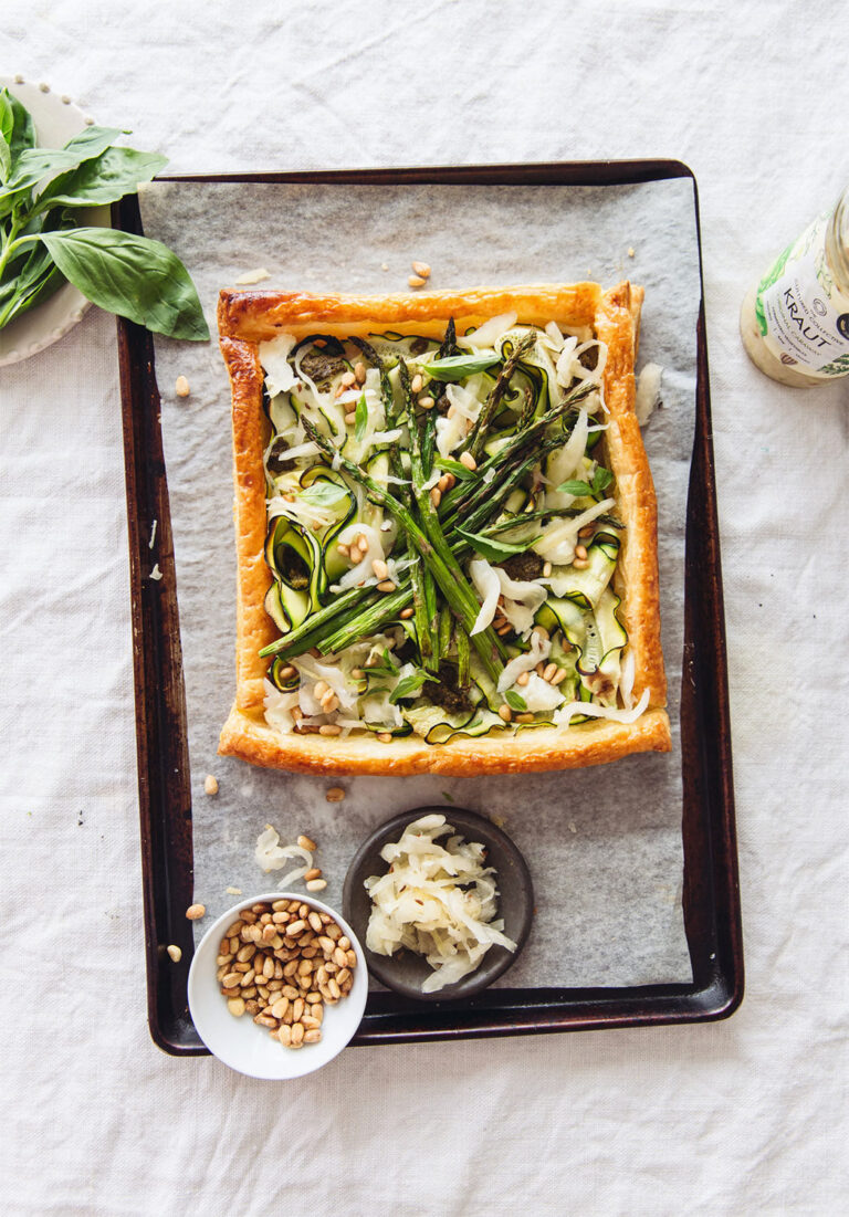 Overhead shot of a rustic tart on a baking sheet, featuring a golden-brown puff pastry crust filled with courgette ribbons, asparagus spears, fresh basil, pine nuts, and sprinkles of sauerkraut; a small bowl of pine nuts and a dish of sauerkraut are beside the tart.