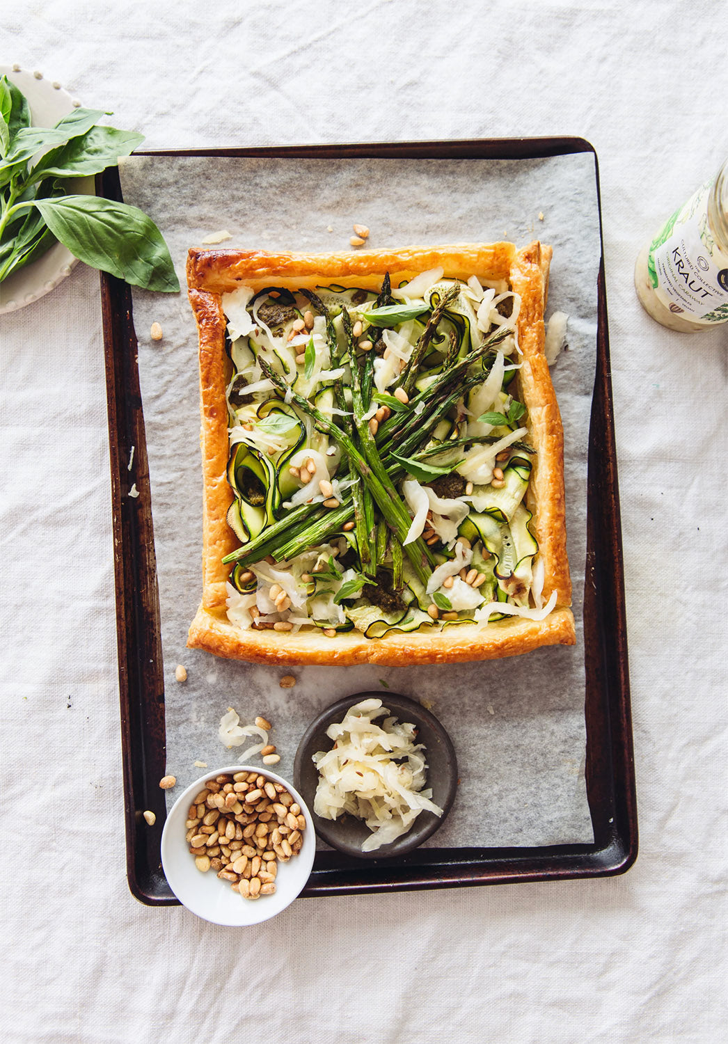 Overhead shot of a rustic tart on a baking sheet, featuring a golden-brown puff pastry crust filled with courgette ribbons, asparagus spears, fresh basil, pine nuts, and sprinkles of sauerkraut; a small bowl of pine nuts and a dish of sauerkraut are beside the tart.