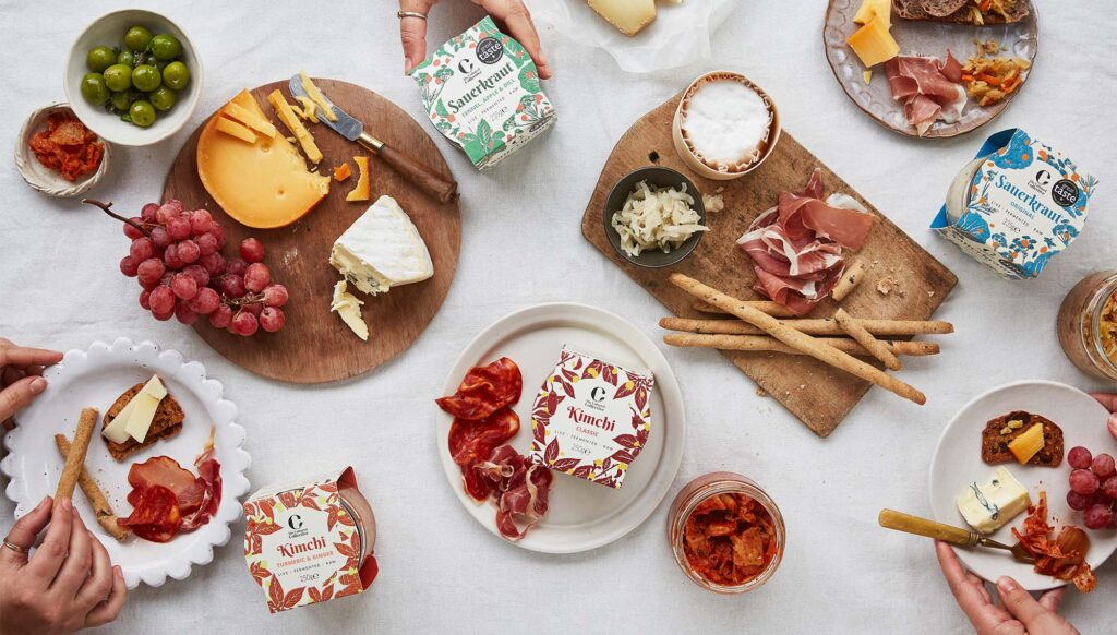 A table set with an assortment of cheeses, cured meats, breadsticks, grapes, olives, and small jars of live fermented kimchi and live sauerkraut. Several hands reach in from the edges of the frame to assemble snacks on plates. Packaged kimchi and sauerkraut containers are scattered among wooden boards holding sliced cheese, prosciutto, and small bowls of fermented vegetables.