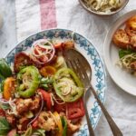 Overhead shot of a colorful panzanella salad on a decorative white and blue plate, with heirloom tomatoes, red onion slices, croutons, capers, and basil, a separate small bowl of sauerkraut, and a small side dish of bread.