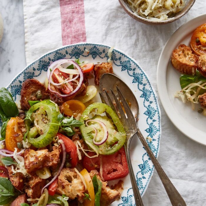 Overhead shot of a colorful panzanella salad on a decorative white and blue plate, with heirloom tomatoes, red onion slices, croutons, capers, and basil, a separate small bowl of sauerkraut, and a small side dish of bread.