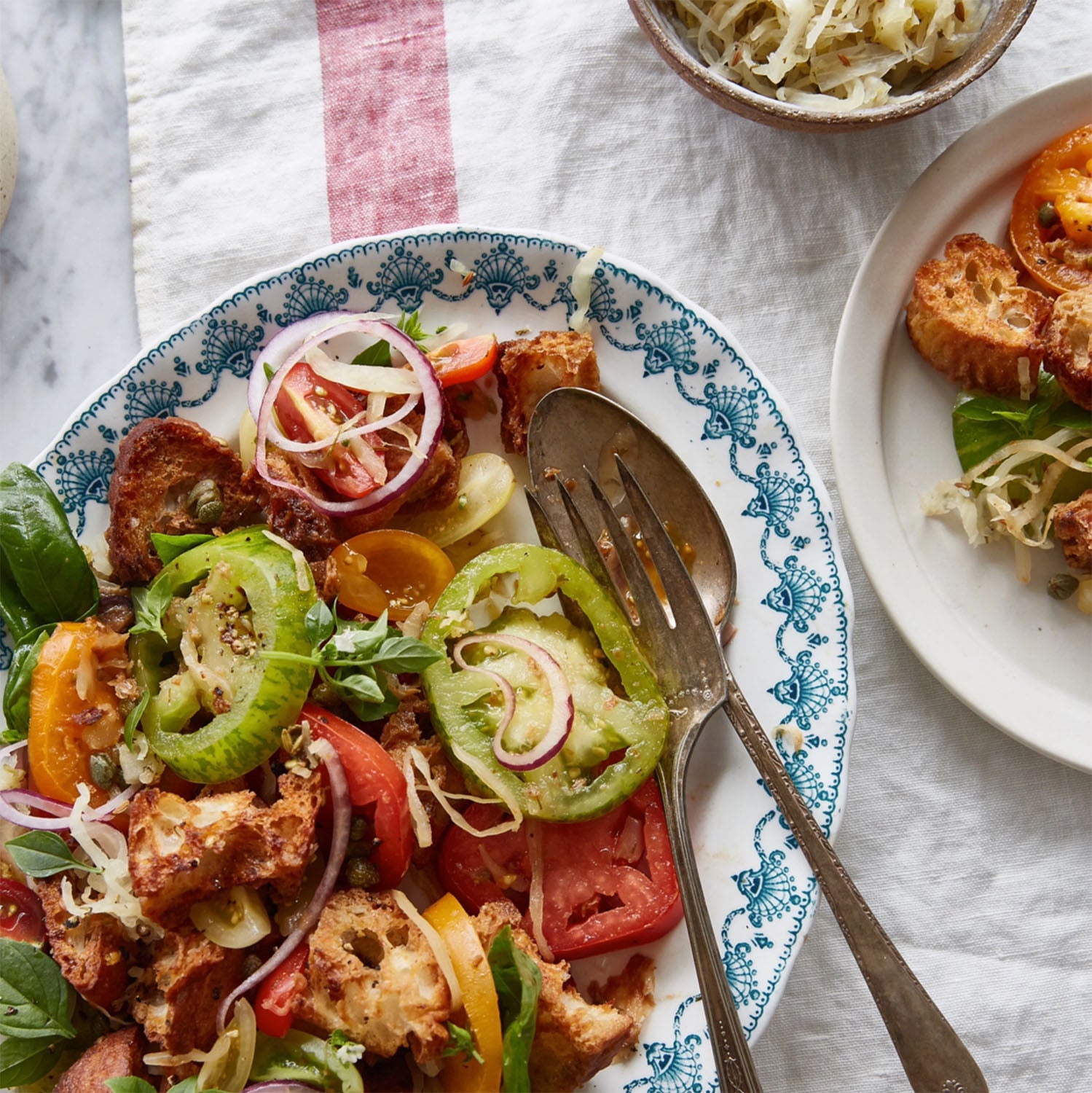 Overhead shot of a colorful panzanella salad on a decorative white and blue plate, with heirloom tomatoes, red onion slices, croutons, capers, and basil, a separate small bowl of sauerkraut, and a small side dish of bread.