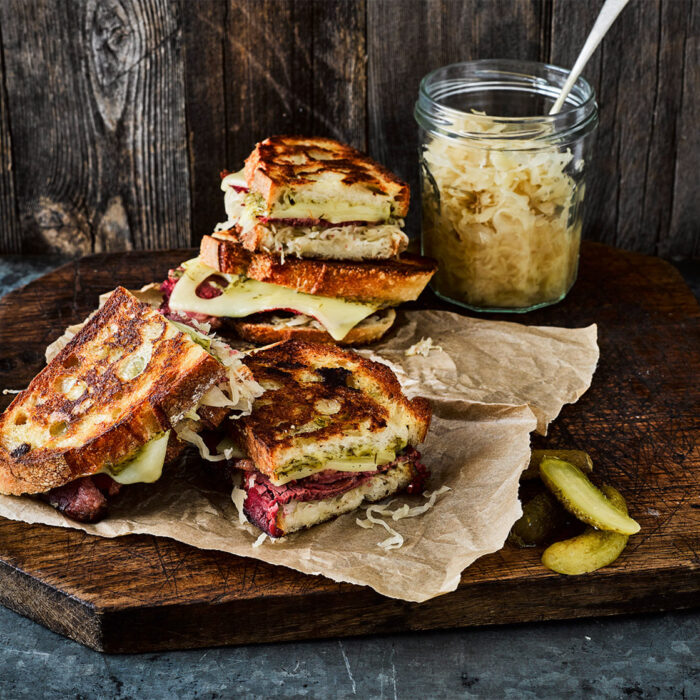 Grilled cheese sandwiches with sourdough bread, Swiss cheese, sauerkraut, and pesto on a wooden board with a jar of sauerkraut and pickle slices.