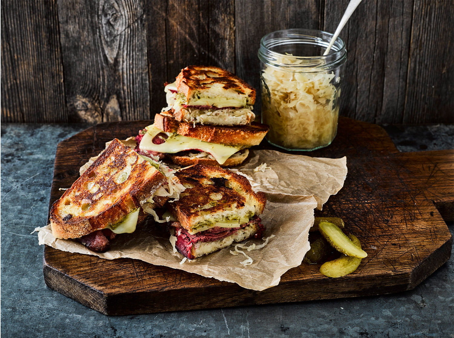 Grilled cheese sandwiches with sourdough bread, Swiss cheese, sauerkraut, and pesto on a wooden board with a jar of sauerkraut and pickle slices.