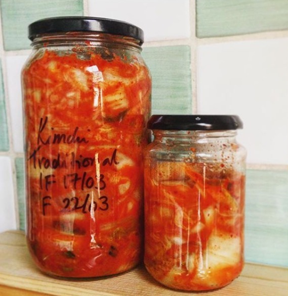 Two glass jars filled with red kimchi on a wooden surface, with a tiled background. The larger jar is labeled "Kimchi Traditional IF 17/03 F 22/03." The kimchi in both jars appears to be a mix of red chili paste and fermented vegetables.