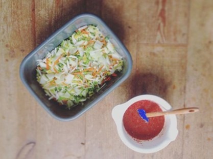 Overhead shot of a meal preparation on a wooden surface. A rectangular container holds mixed chopped vegetables, and a small bowl contains a red sauce with a blue spatula and a wooden spoon resting inside.