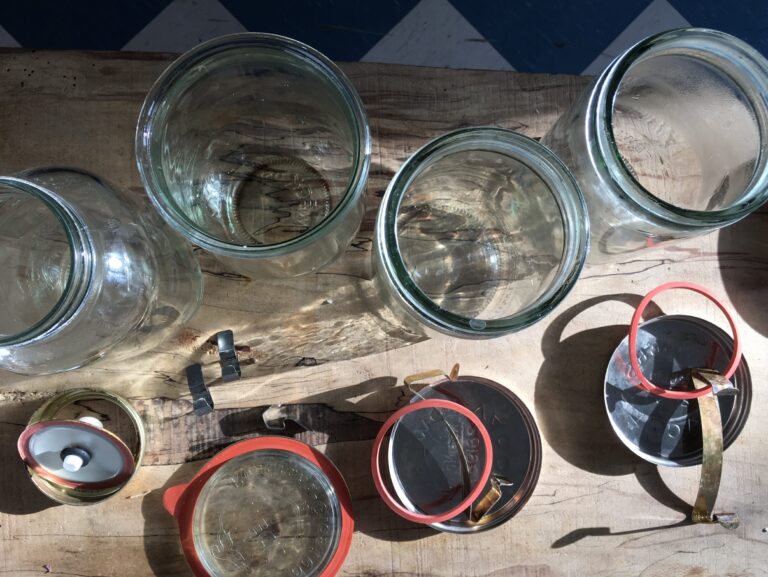 Overhead shot of glass jars, lids, and sealing rings arranged on a wooden surface, suggesting canning preparations; several empty glass jars are visible with open mouths, surrounded by lids, orange rubber rings, and metal closure clips.