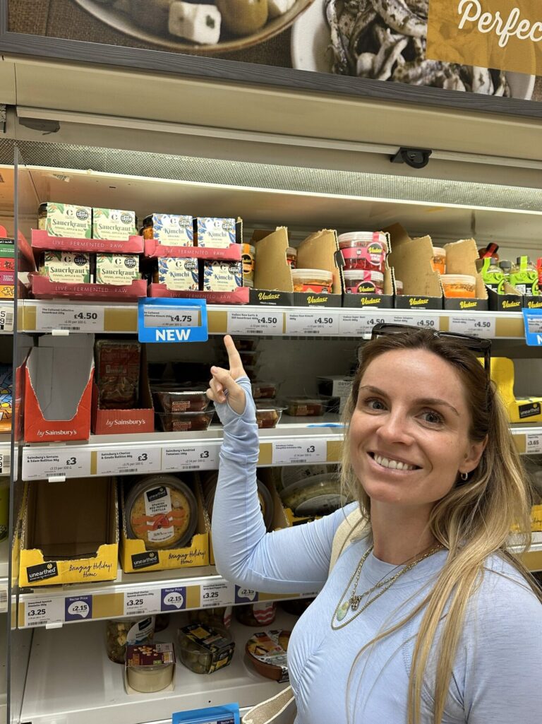 Founder Nikki Peters, a woman with long blonde hair smiles while pointing towards a "NEW" sign on a shelf in a grocery store, with various jars and containers of fermented foods like kimchi and sauerkraut visible.