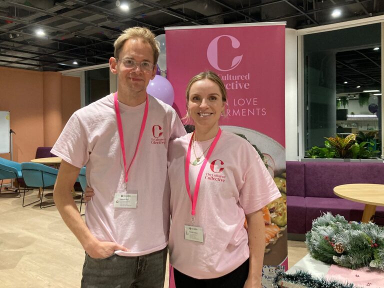 Two people wearing pink t-shirts with "The Cultured Collective" logo and lanyards, standing in front of a banner with the same logo and the words "Love Ferments." They are in a modern event space, with a purple sofa, round tables, and decorative elements.
