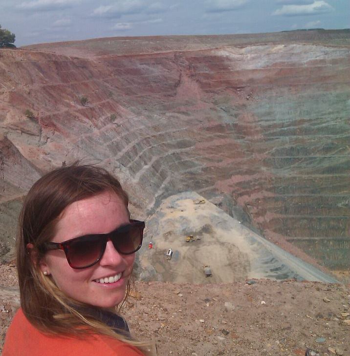 Smiling woman wearing sunglasses in front of a large, terraced open-pit mine, likely a copper mine, with heavy machinery visible on a central mound.