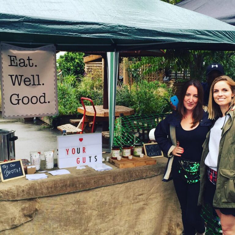 Two women standing at a food market stall advertising "Eat. Well. Good." and "Love Your Guts", with jars of fermented foods and a sign for broth. The stall is under a green marquee tent with a garden in the background.