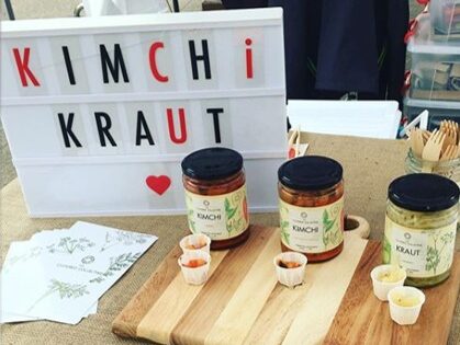 Display of homemade kimchi and kraut at a market stall, with jars of the fermented food on a wooden cutting board, samples in paper cups, and a lighted sign that says "Kimchi Kraut" with a heart icon.