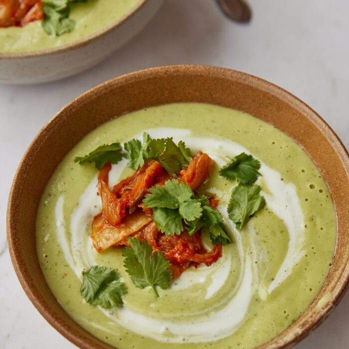 A close-up shot of a bowl of green soup garnished with red kimchi, fresh coriander, and a swirl of cream, set on a white marble surface next to a second bowl of soup.
