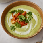 A close-up shot of a bowl of green soup garnished with red kimchi, fresh coriander, and a swirl of cream, set on a white marble surface next to a second bowl of soup.