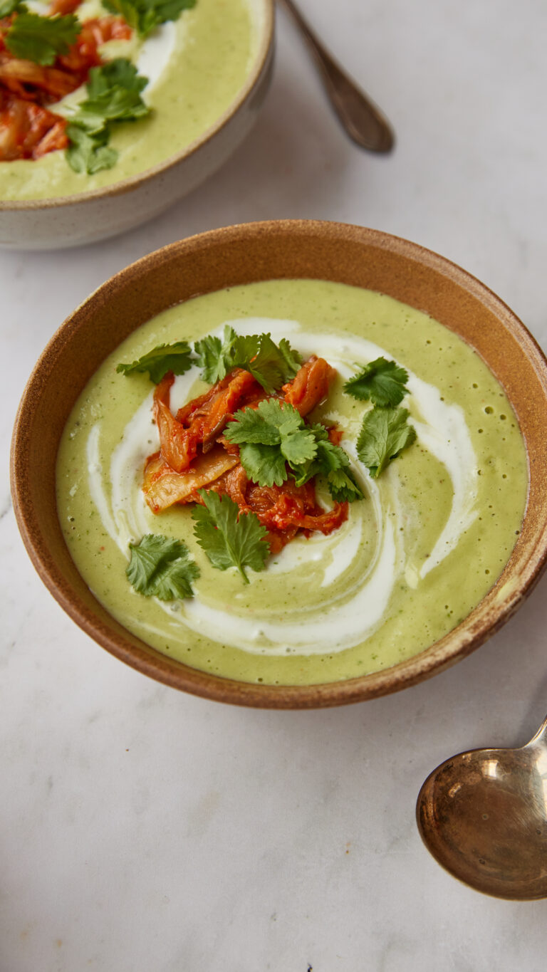 A close-up shot of a bowl of green soup garnished with red kimchi, fresh coriander, and a swirl of cream, set on a white marble surface next to a second bowl of soup.