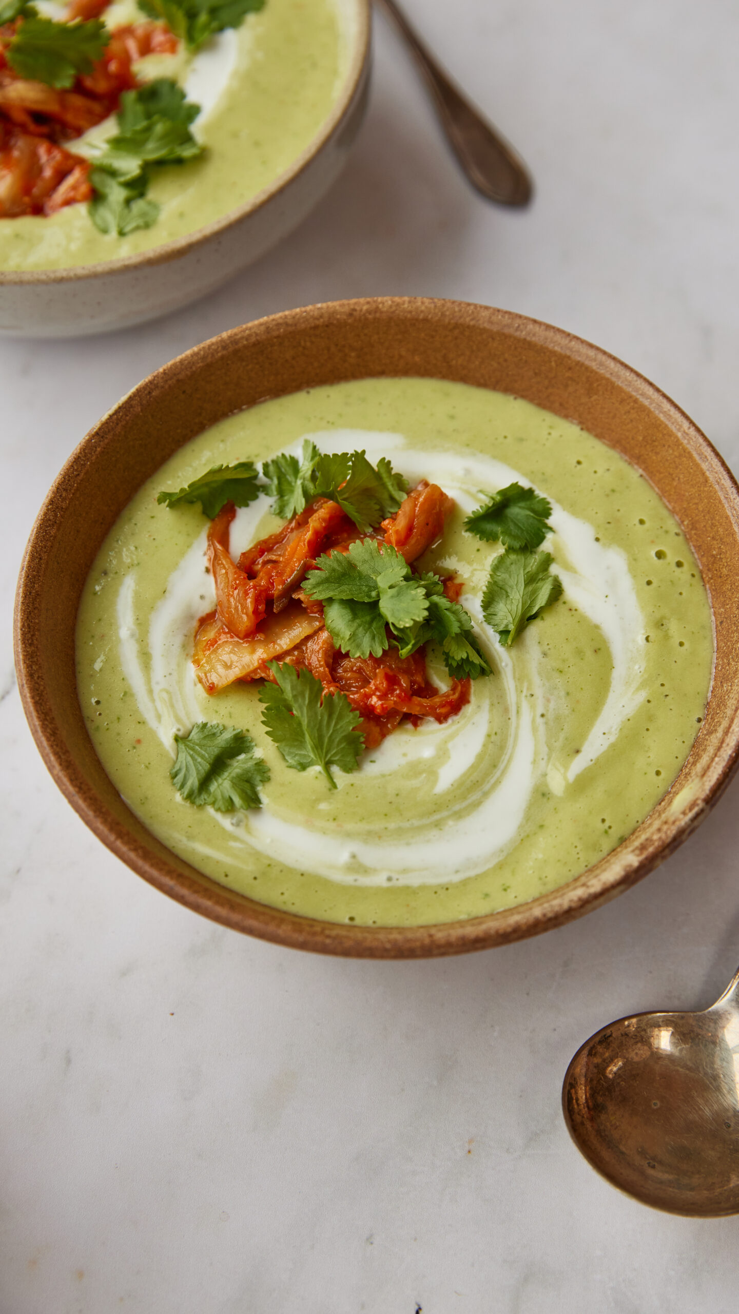 A close-up shot of a bowl of green soup garnished with red kimchi, fresh coriander, and a swirl of cream, set on a white marble surface next to a second bowl of soup.