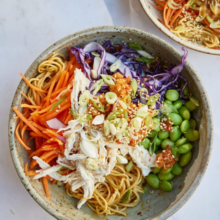 A bowl of Asian-inspired noodles with shredded chicken, edamame, and vegetables on a marble surface.