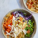 Two bowls of Asian-inspired noodles with shredded chicken, edamame, and vegetables on a marble surface, with a glass of peanut sauce in the background.
