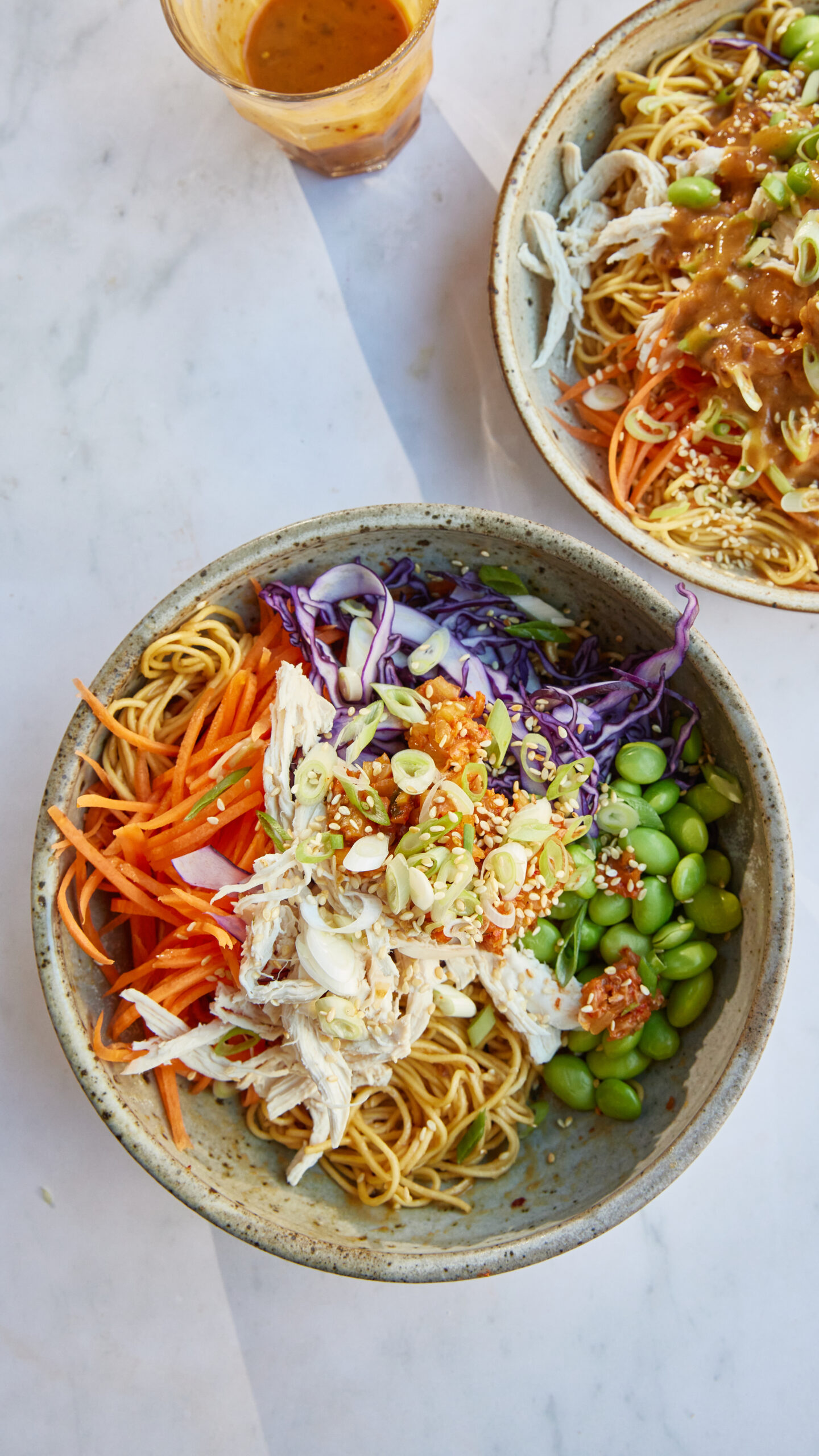 Two bowls of Asian-inspired noodles with shredded chicken, edamame, and vegetables on a marble surface, with a glass of peanut sauce in the background.