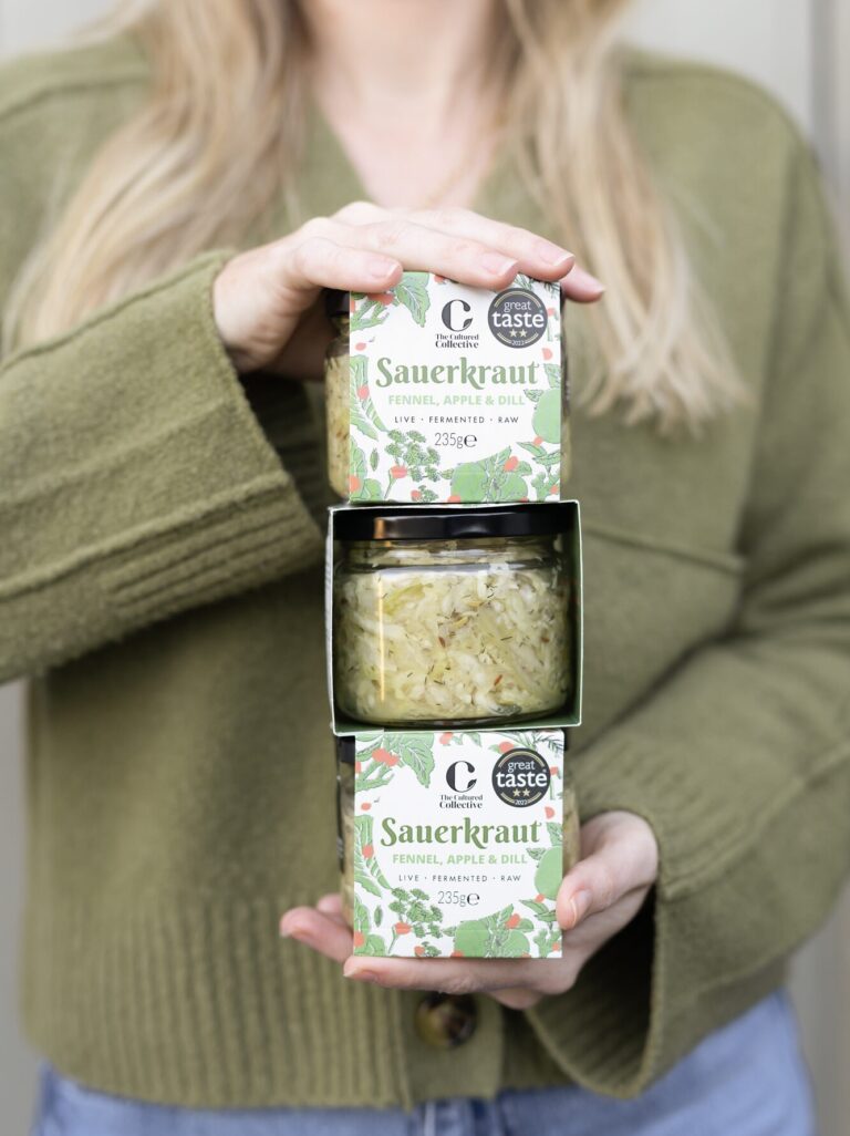 Woman holding a stack of three jars of "The Cultured Collective" Sauerkraut, flavoured with fennel, apple, and dill. Each jar is encased in a decorative green box, and the middle jar is fully visible, displaying the fermented cabbage inside. The boxes feature a "Great Taste" award seal. The woman wears a green sweater.