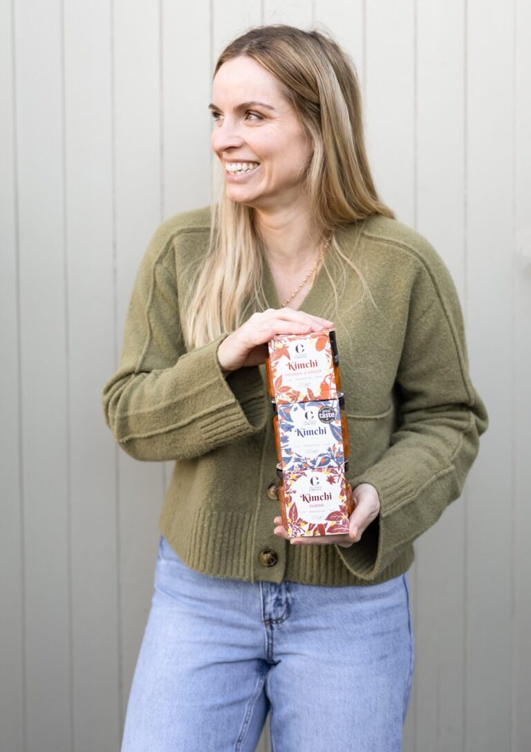 Founder Nikki Peters, a blonde woman smiling and holding a stack of three boxes of "The Cultured Collective Kimchi", with flavors "Turmeric & Ginger," "Classic," and "Original", wearing a green cardigan and jeans.