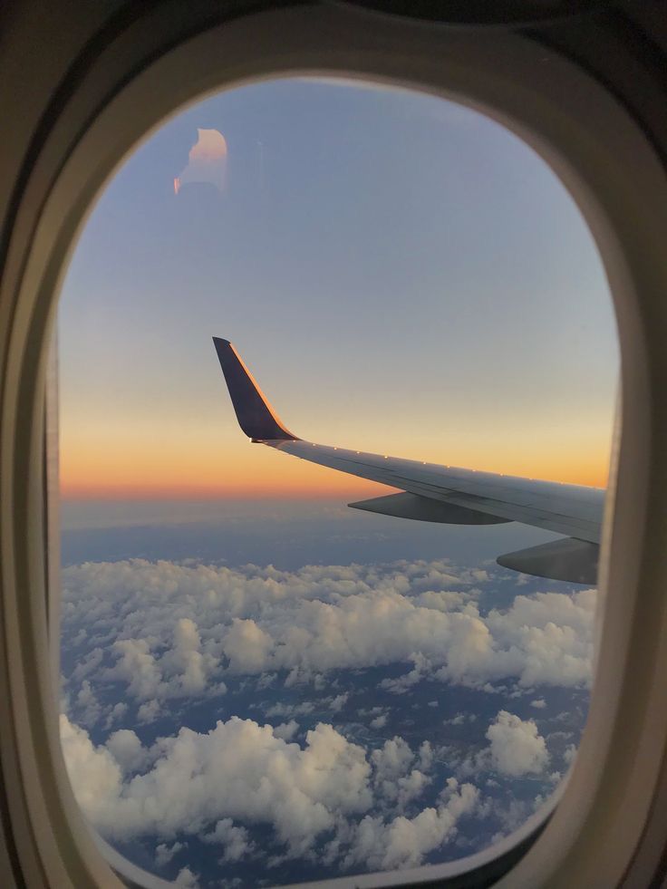 View from an airplane window showcasing a wing silhouetted against a vibrant sunset sky. Below, a thick layer of white clouds is visible, and the frame of the window is dark.