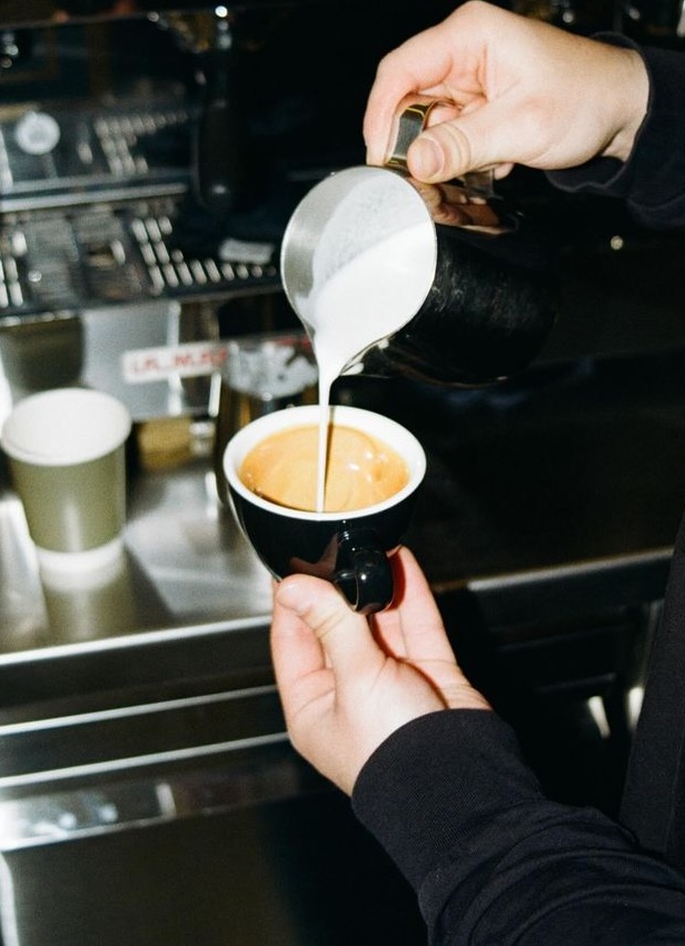 Barista pouring steamed milk from a metal pitcher into a black espresso cup, the cup held in another hand. The cup is filled with coffee, milk is being poured into the center of the cup. A green coffee cup and an espresso machine is visible in the background.