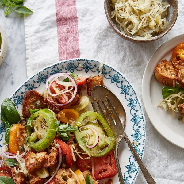 Overhead shot of a colorful panzanella salad on a decorative white and blue plate, with heirloom tomatoes, red onion slices, croutons, capers, and basil, a separate small bowl of sauerkraut, and a small side dish of bread.
