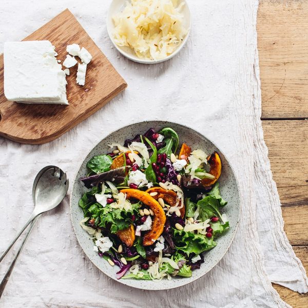 Overhead shot of a colorful salad in a speckled gray bowl, with roasted butternut squash, pomegranate seeds, pine nuts, greens, sauerkraut and crumbled feta cheese, with a wooden cutting board holding a block of feta and a small bowl of sauerkraut nearby.